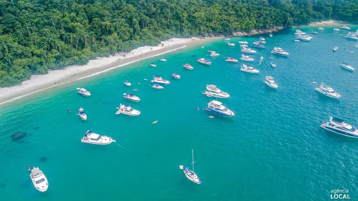 Praia de areia branca entre a mata e o mar turquesa, com dezenas de lanchas ancoradas