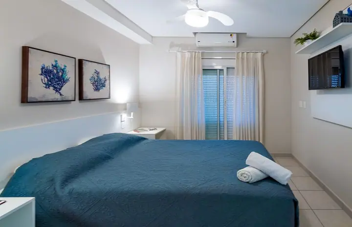 Quarto de apartamento com colcha azul-petróleo, quadros de corais, ventilador de teto e porta de varanda