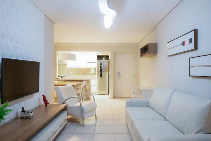 Sala de apartamento com sofá branco, poltrona, TV, quadros com remos e cozinha integrada ao fundo