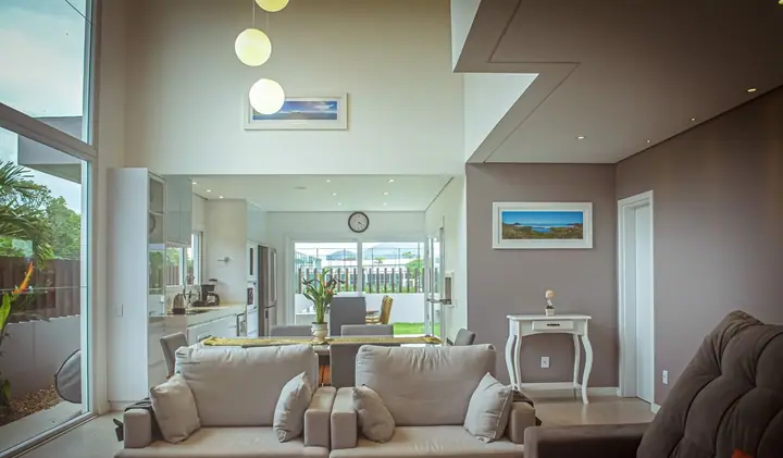 Living de casa contemporânea com pé-direito duplo, sofás claros, pendentes de vidro e cozinha integrada voltada para o jardim