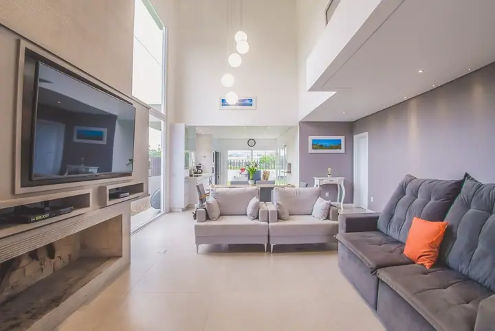 Sala de TV com sofá cinza, almofada laranja, painel de TV e pé-direito duplo com mezanino