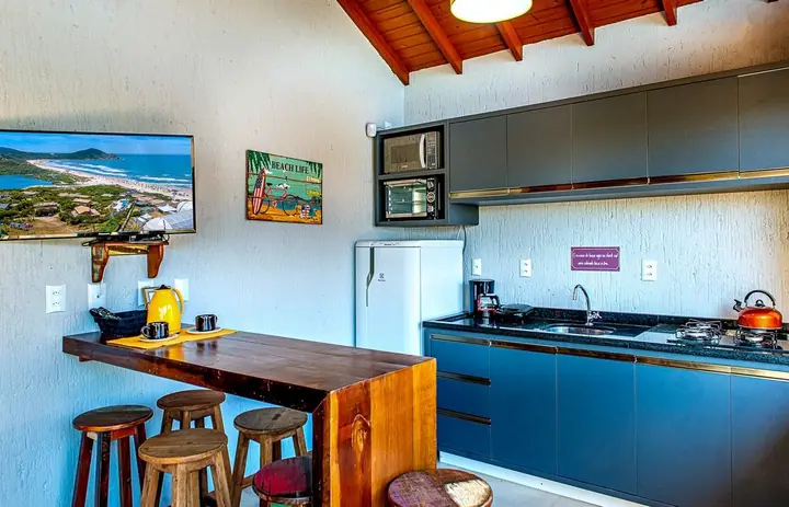 Cozinha de chalé com forro de madeira, armários escuros, bancada de madeira maciça, banquetas e quadros de praia