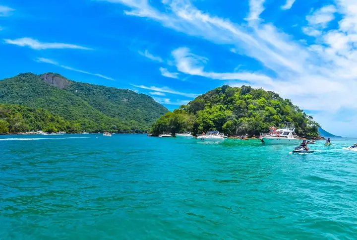 Ilha coberta de mata em mar azul, com lanchas ancoradas e morros ao fundo