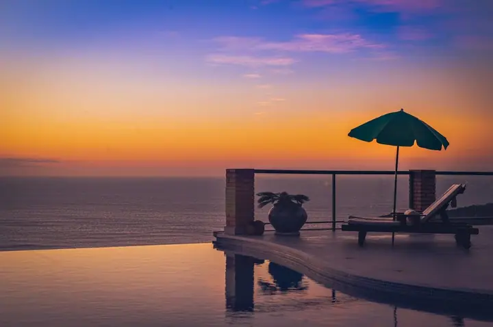 Piscina de borda infinita refletindo o céu do entardecer, com guarda-sol, espreguiçadeira e o mar ao fundo