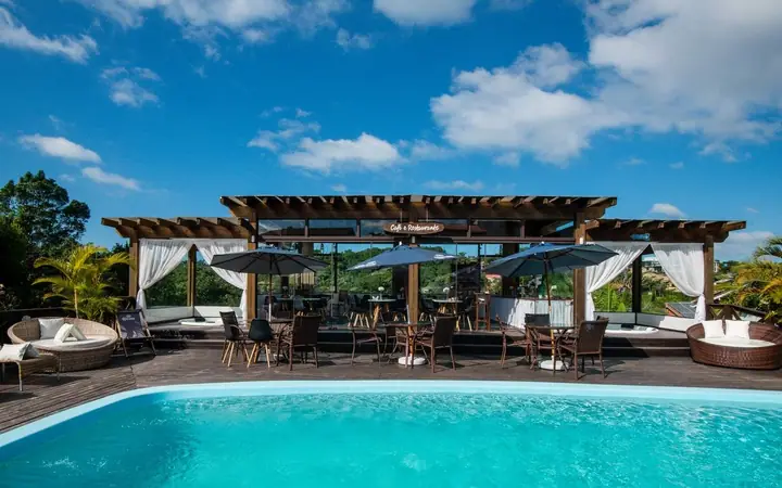 Piscina de pousada com deck, lounge sob pergolado com cortinas brancas, mesas e guarda-sóis, sob céu azul