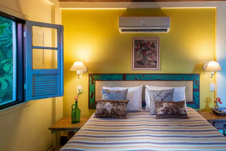 Quarto de pousada com parede amarela, janela de madeira azul aberta, cama de casal com colcha listrada e quadro de flores