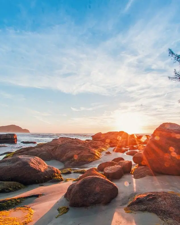 Sol baixo entre as pedras da praia, com árvore no costão e céu azul com nuvens