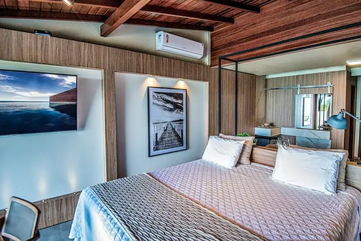 Suíte com forro de madeira, cama de casal com colcha matelassê, quadro de píer, TV e box de vidro ao fundo